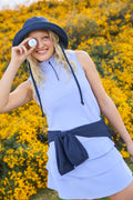 Woman in a stardust sleeveless top and skirt with navy belt and hat, standing in front of yellow flowers.