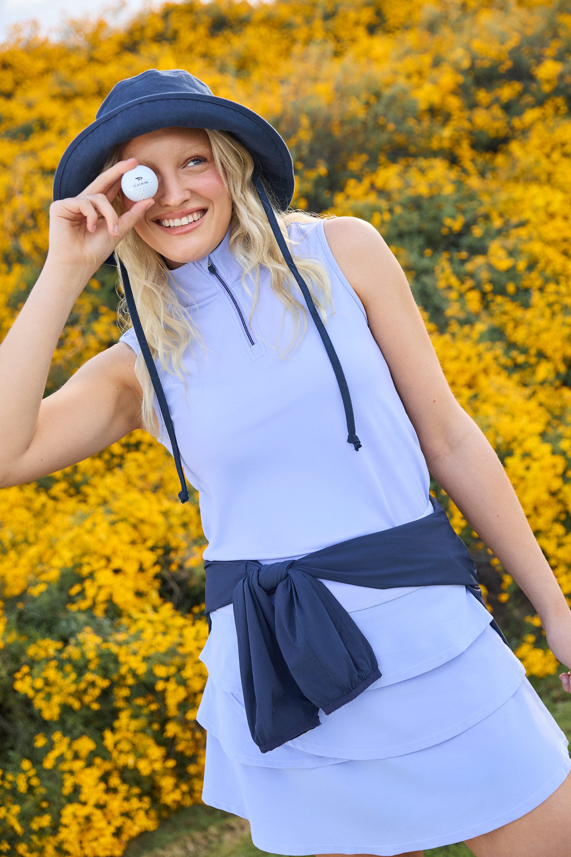 Woman in a stardust sleeveless top and skirt with navy belt and hat, standing in front of yellow flowers.