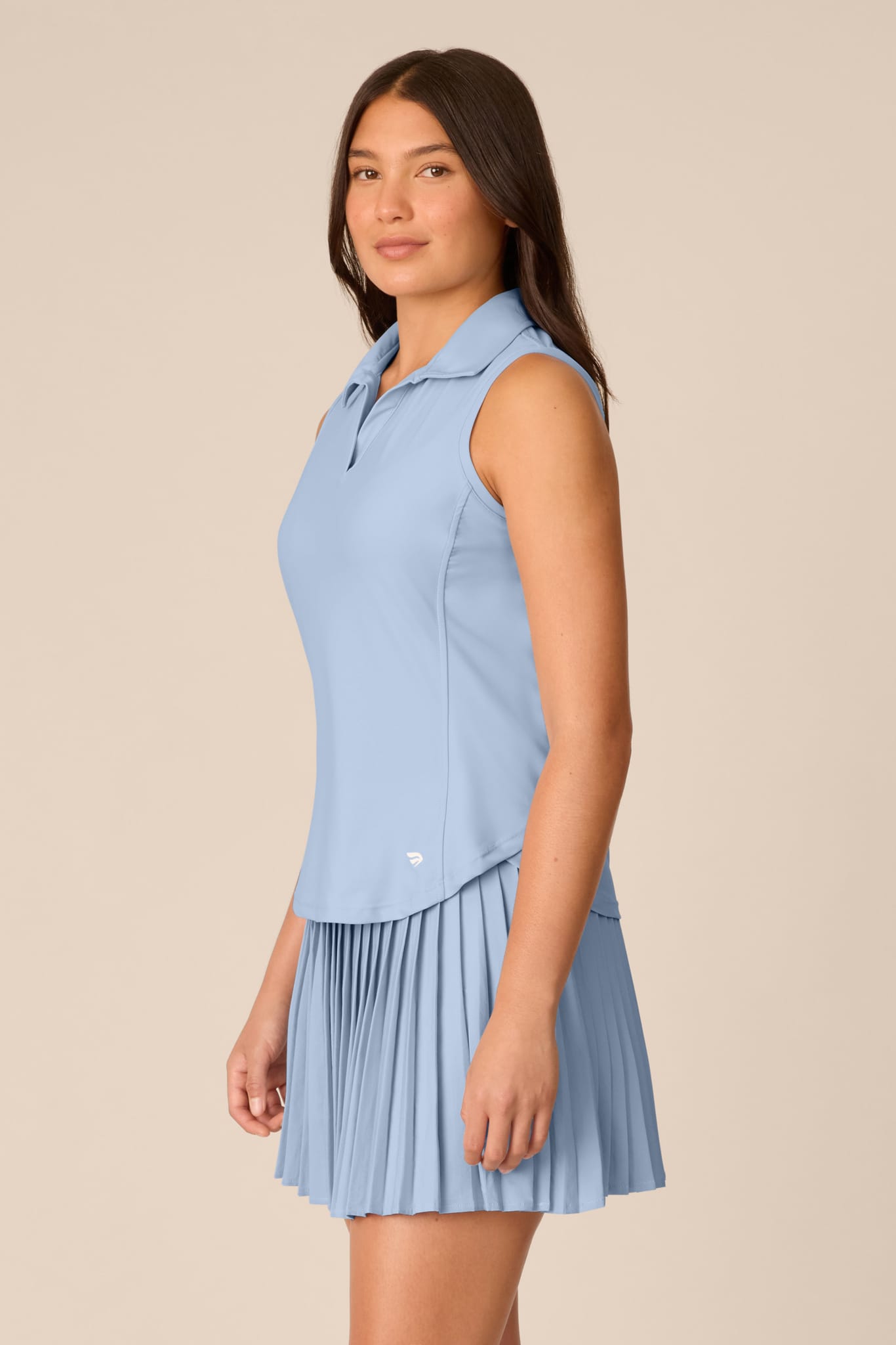 Skyline | A woman modeling a light blue sleeveless polo shirt with a matching pleated mini skirt against a solid beige background.