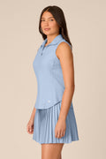 Skyline | A woman modeling a light blue sleeveless polo shirt with a matching pleated mini skirt against a solid beige background.