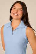 Skyline | A smiling young woman with long dark hair wearing a light blue sleeveless polo shirt, photographed against a plain warm beige studio background.
