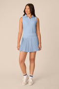 Skyline | A woman modeling a sleeveless light blue polo dress with a pleated skirt, paired with white socks and white sneakers, against a solid beige studio background.