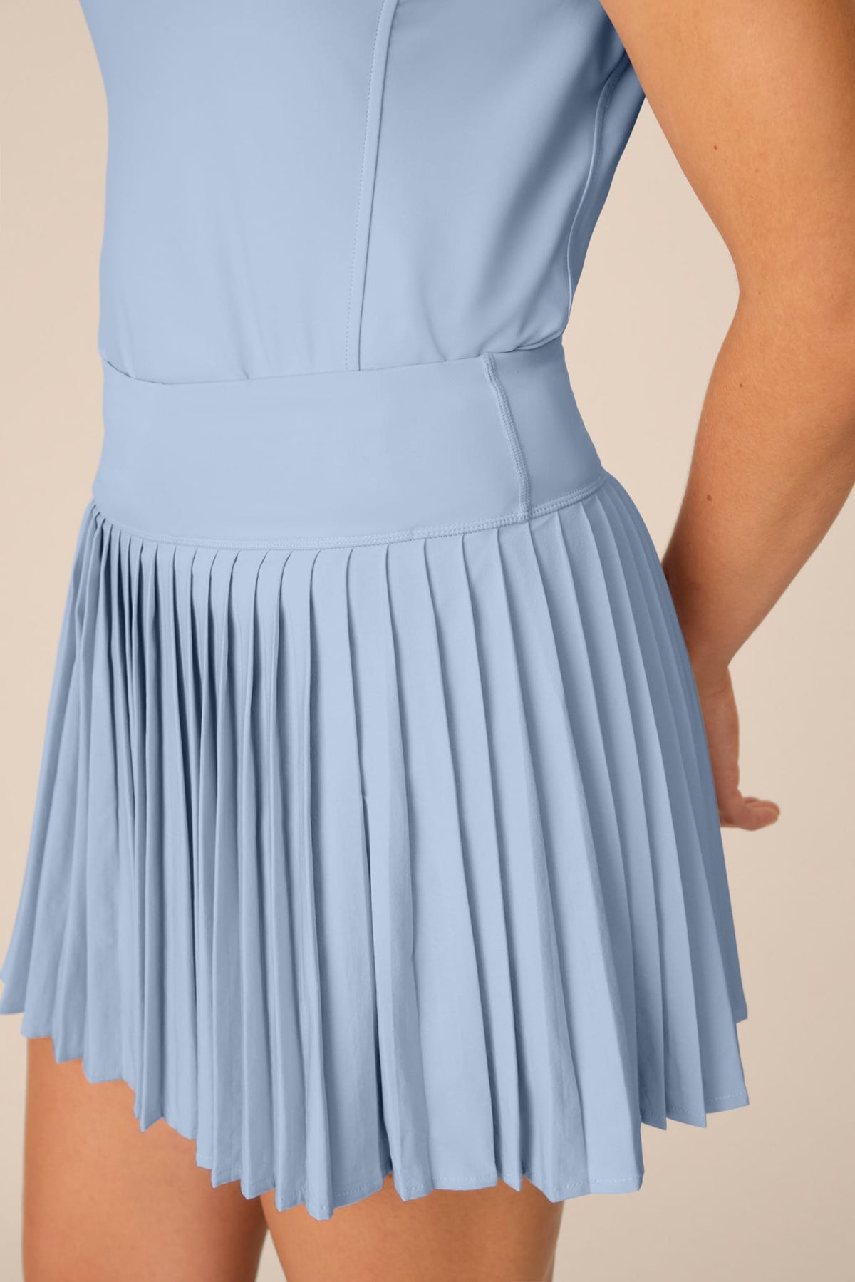 Skyline | A close-up of a light blue pleated tennis skirt worn by a model, showing the pleated texture and waistband against a solid beige background.