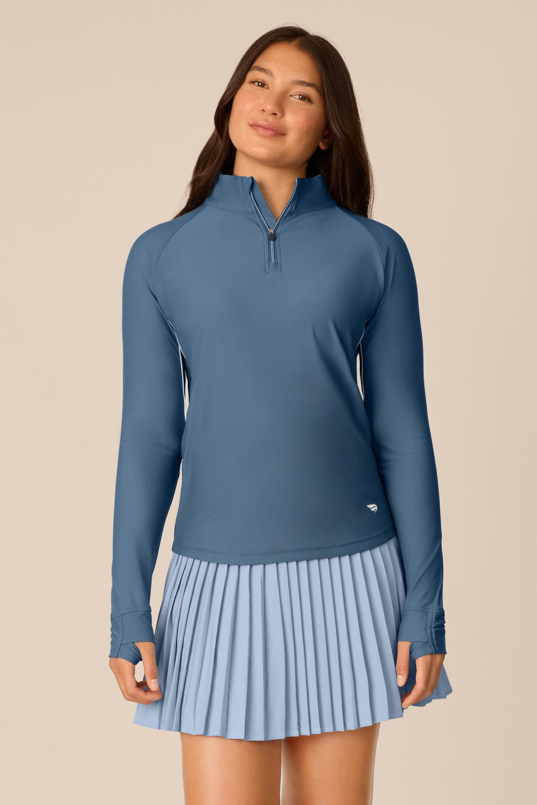 Slate | A woman modeling a slate blue long-sleeve athletic top and matching pleated skirt set against a solid beige background.