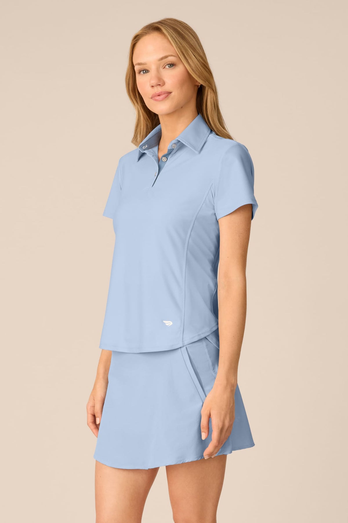 Skyline | A woman modeling a light blue short-sleeve collared polo shirt and matching high-waisted athletic skirt against a solid beige studio background.