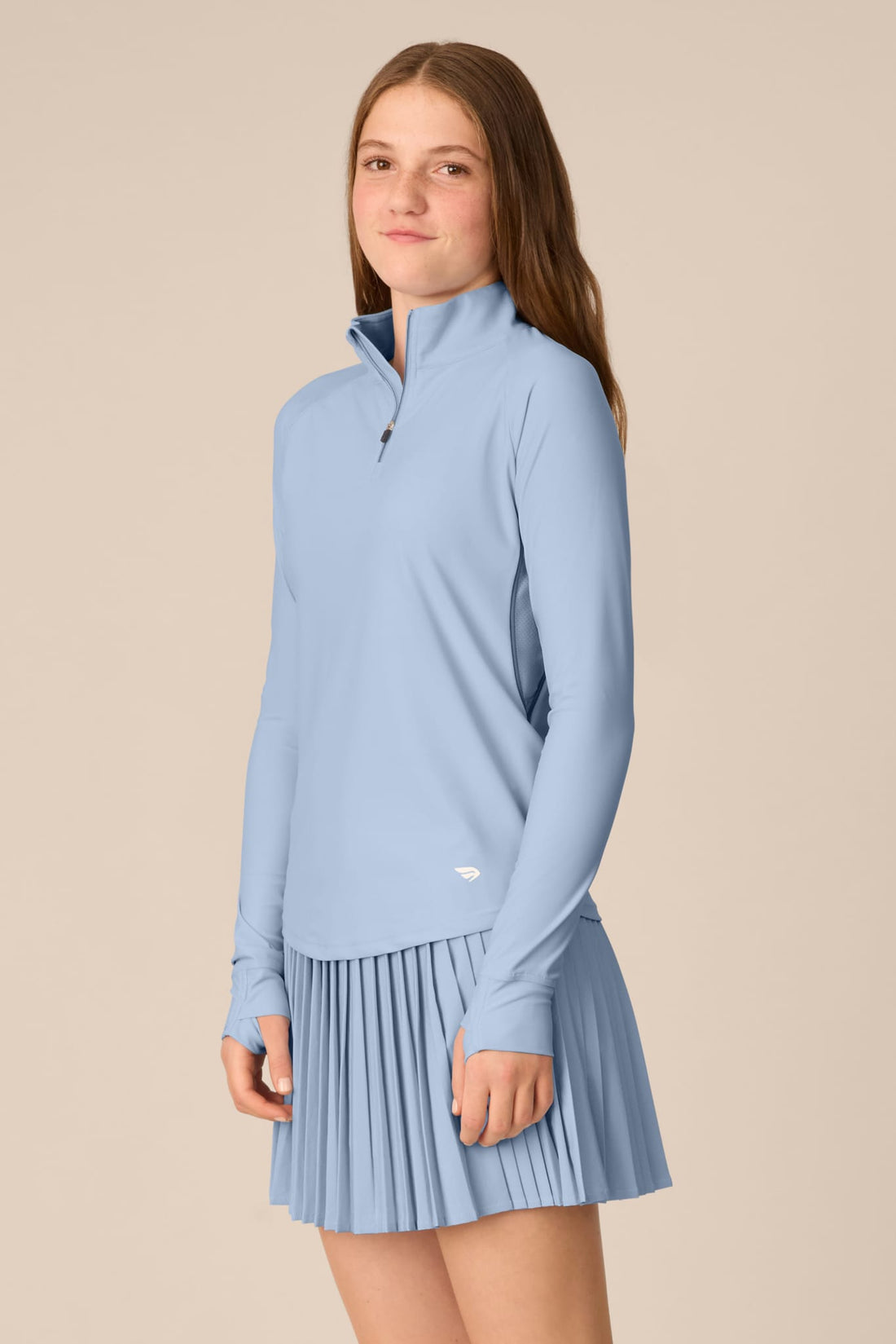 Skyline | A young woman modeling a light blue athletic long-sleeve quarter-zip top and matching pleated skirt against a solid beige background.