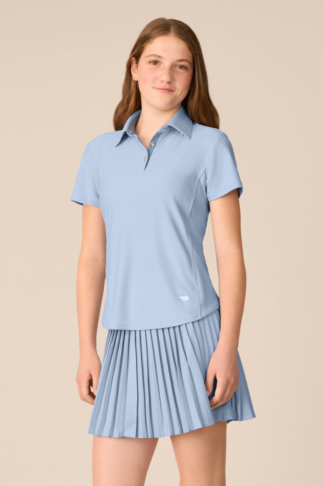 Skyline | A young woman modeling a light blue short-sleeve polo shirt and matching pleated tennis skirt against a solid beige background.