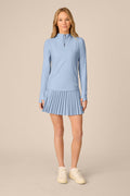 Skyline | A woman modeling a light blue long-sleeve athletic top and matching pleated mini skirt set, paired with white sneakers, against a solid beige studio background.