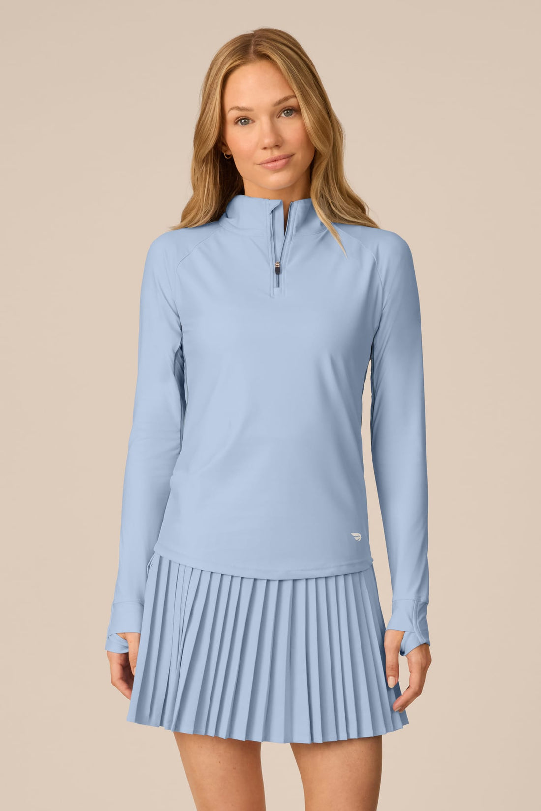 Skyline | A woman modeling a light blue long-sleeve athletic top and matching pleated skirt set against a solid beige background.