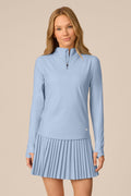 Skyline | A woman modeling a light blue long-sleeve athletic top and matching pleated skirt set against a solid beige background.