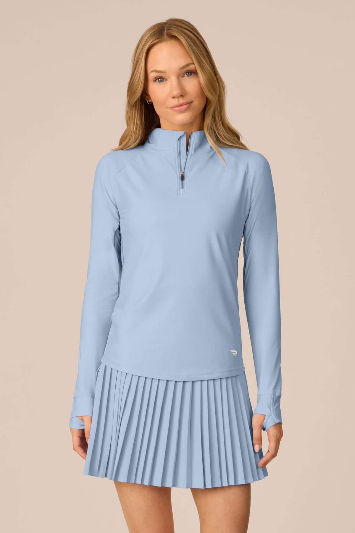 Skyline | A woman modeling a light blue long-sleeve athletic top and matching pleated skirt set against a solid beige background.