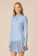 Skyline | A woman modeling a light blue long-sleeve athletic top and matching pleated mini skirt against a solid beige studio background.