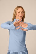 Skyline | A woman modeling a light blue long-sleeve athletic top with a mock neck and matching pleated skirt against a solid beige background.