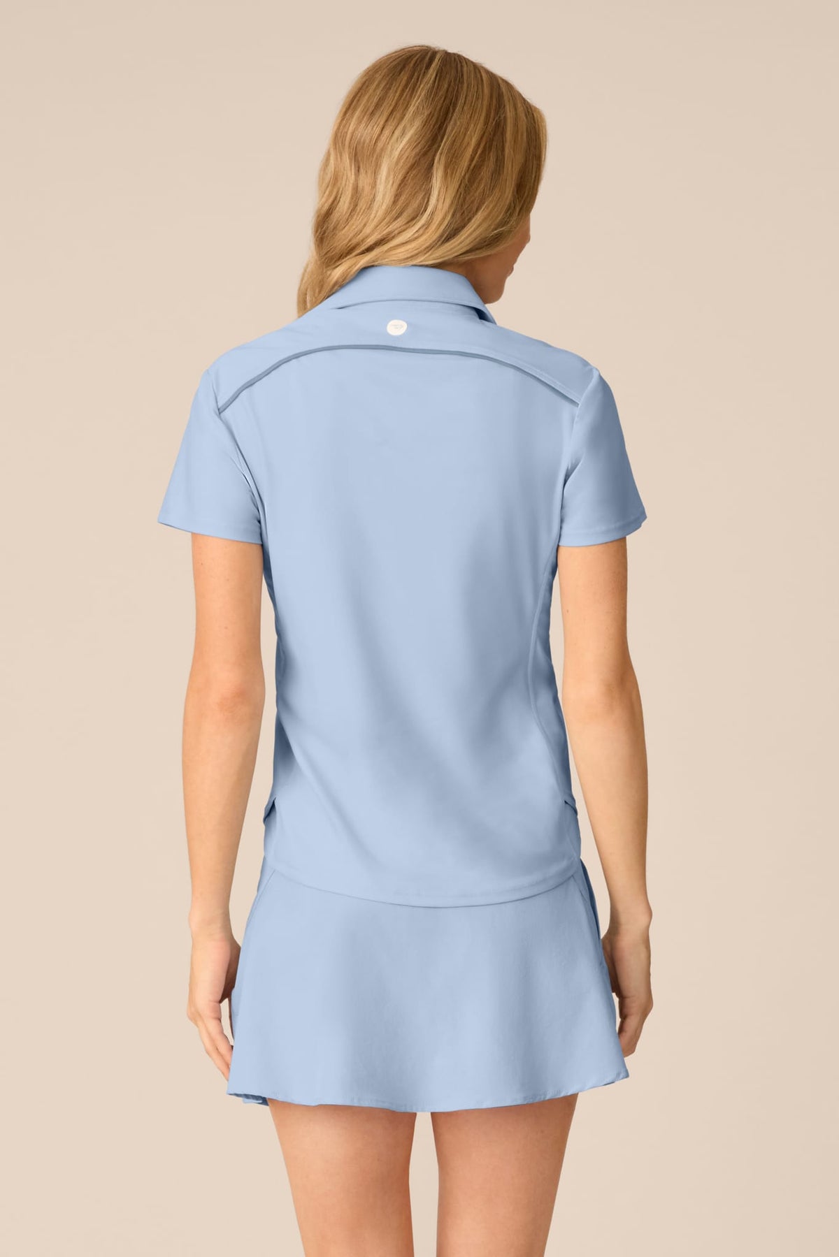 Skyline | A woman viewed from the back wearing a light blue short-sleeve collared polo-style top and matching mini skirt against a plain beige studio background.
