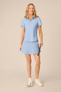 Skyline | A woman modeling a light blue short-sleeve polo shirt and matching skirt set, holding a golf club against a plain beige studio background.