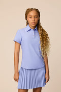 Stardust | Junior golfer wearing a Stardust color Lexi polo shirt and pleated skirt on a beige background