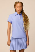 Stardust | Junior golfer wearing a Stardust color Lexi polo shirt and pleated skirt on a beige background