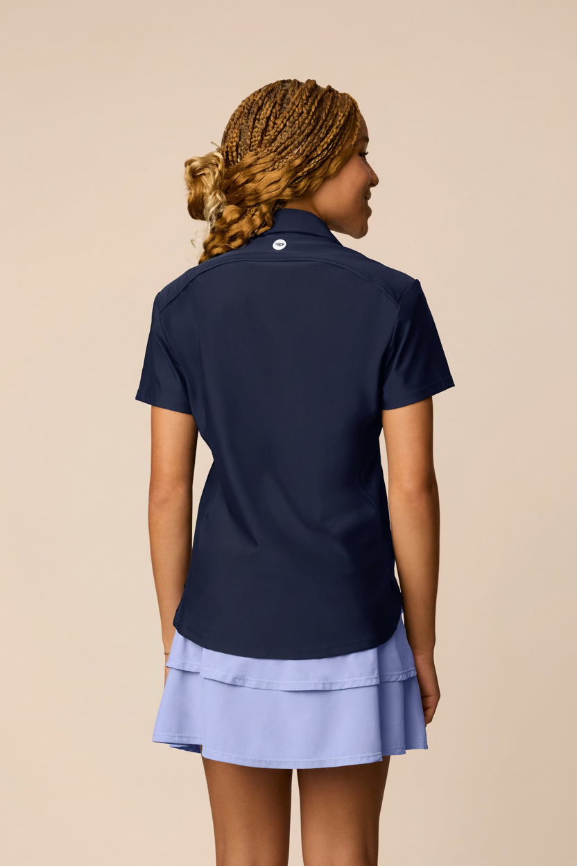 Midnight | Junior golfer wearing a Midnight color Lexi polo shirt and pleated skirt on a beige background