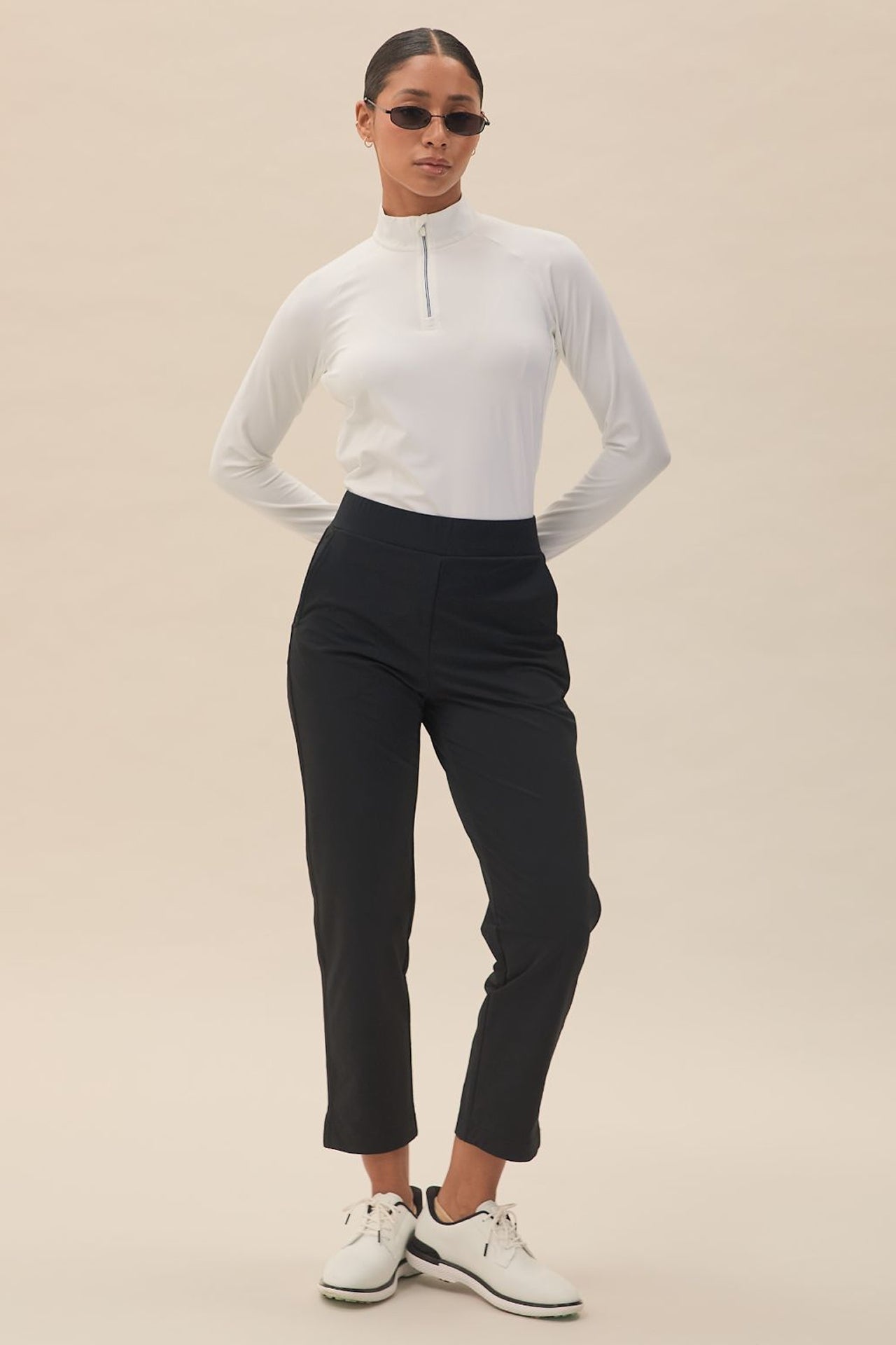 Black | A woman is standing, wearing black slim-fit mid-waist pants with a white built-in tee holder on the back waistband. She has on a white long-sleeve top, sunglasses, and white shoes.
