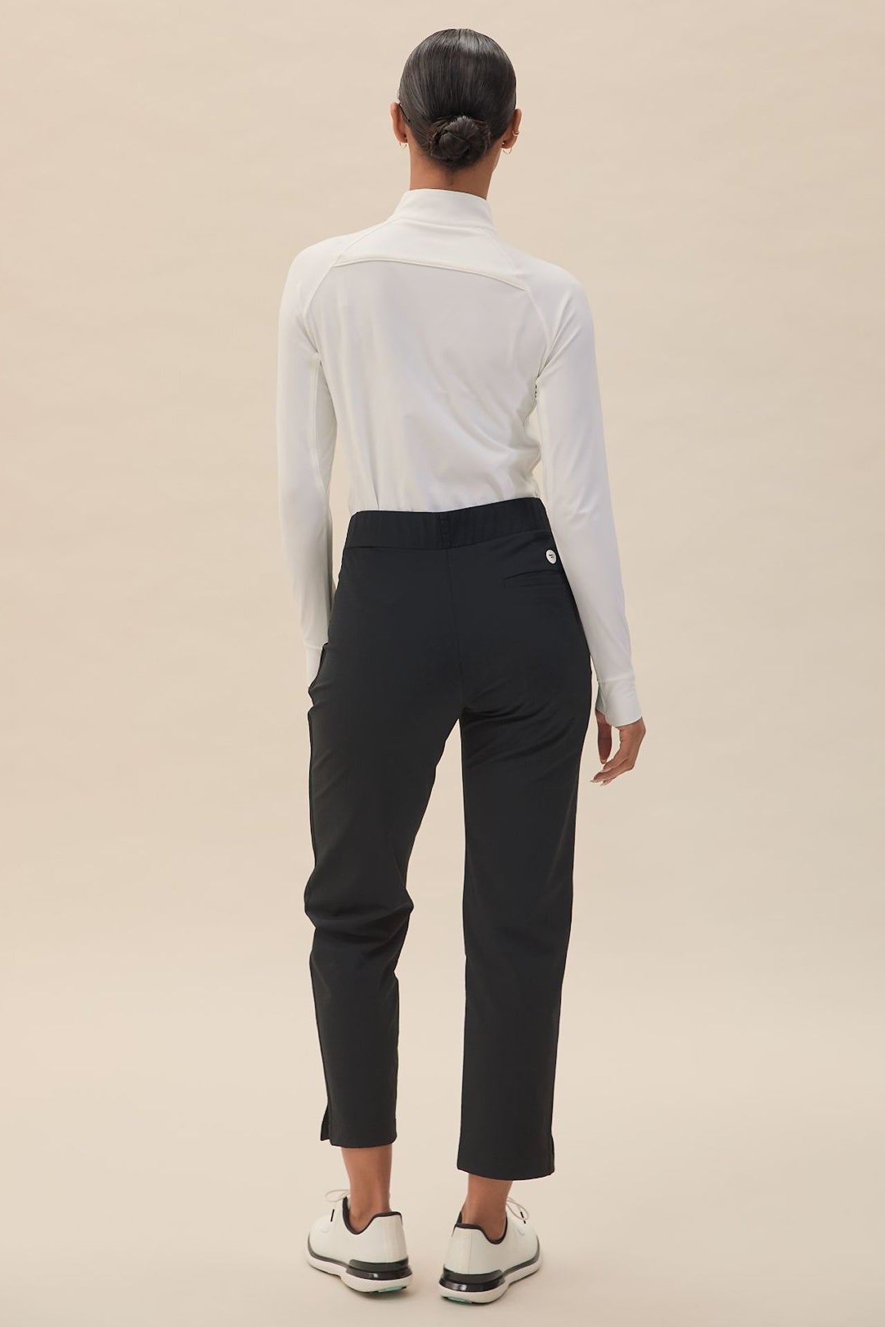 Black | A woman is standing, wearing black slim-fit mid-waist pants with a white built-in tee holder on the back waistband. She has on a white long-sleeve top, sunglasses, and white shoes.