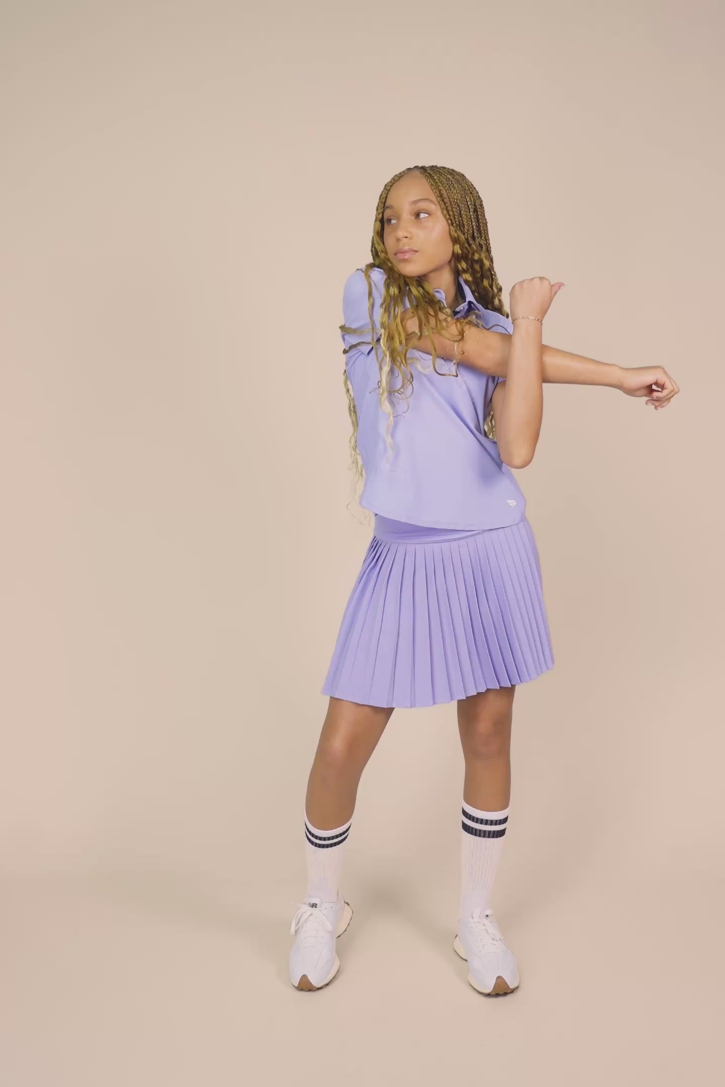 Stardust | Junior golfer wearing a Stardust color Lexi polo shirt and pleated skirt on a beige background